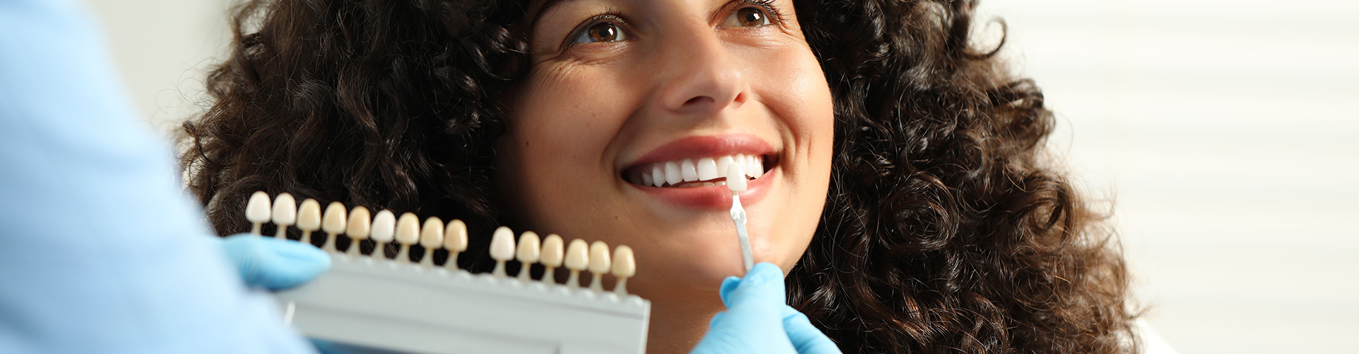 Gloved hand holding tooth shade-matching tool next to woman's smile.